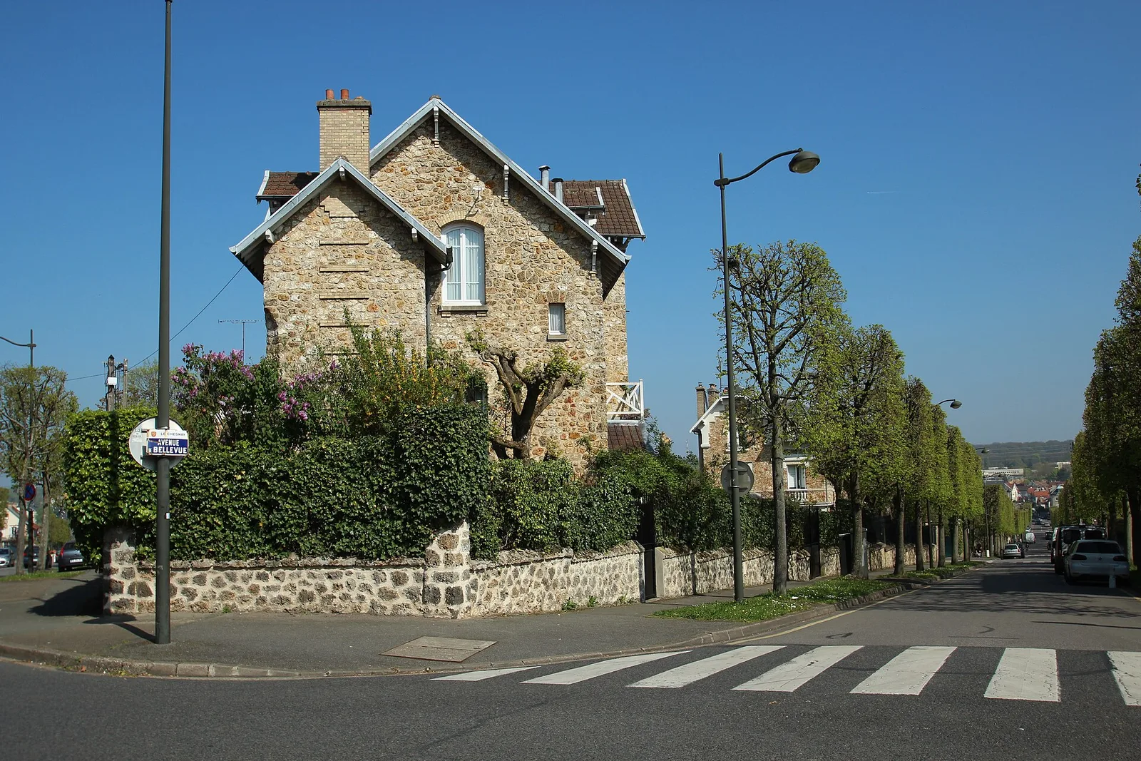 Maison en pierre meulière à pignons avec jardin clos, vue depuis un carrefour résidentiel