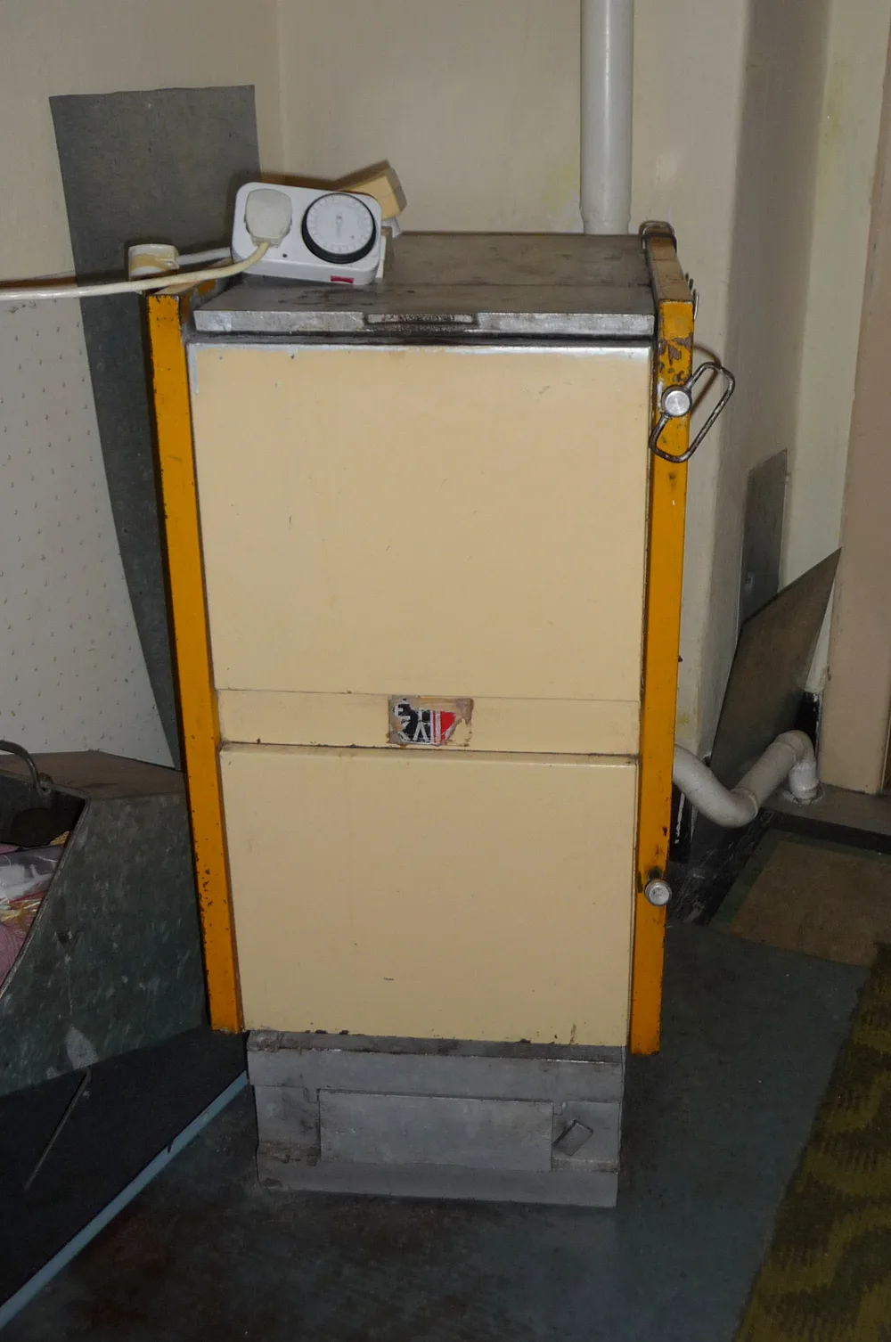 Ancienne chaudière à combustible solide à chargement par le haut, carrosserie beige et cadre orange, installée dans une pièce d'habitation avec son conduit de fumée