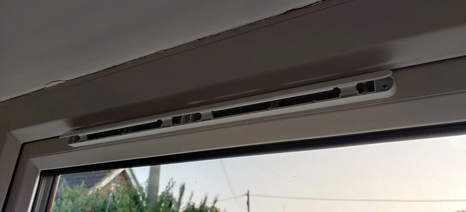 Entrée d'air blanche en réglette fixée sur la traverse haute d'une fenêtre en PVC, vue de l'intérieur, avec le jardin visible à travers le vitrage