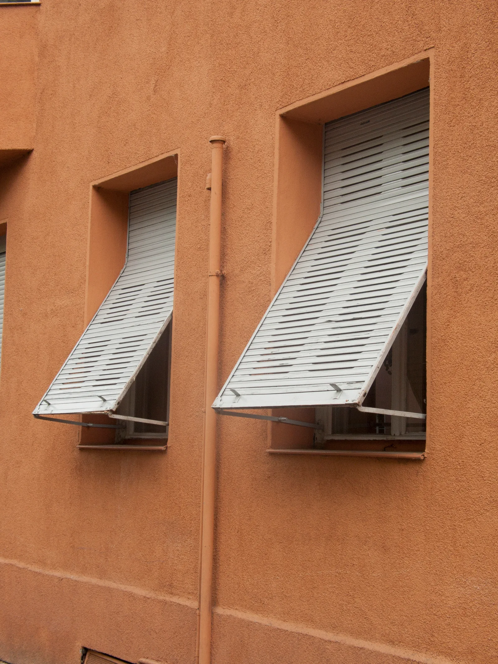 Deux fenêtres d'un immeuble crépi équipées de volets extérieurs à lames blanches relevés en position inclinée, en projection devant la vitre