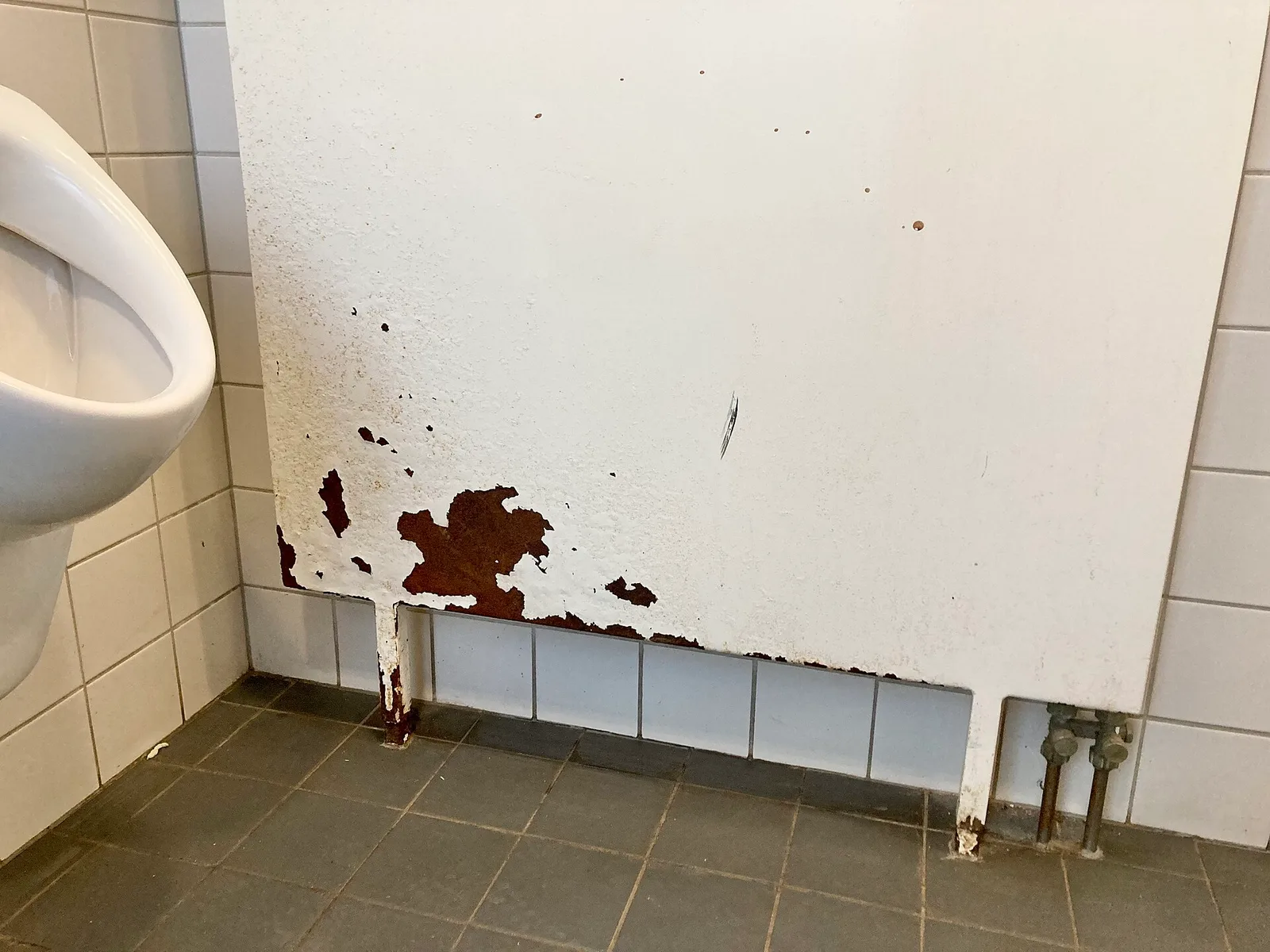 Radiateur à panneaux blanc fixé au mur d'un local carrelé, dont la peinture a éclaté sur le bas, laissant apparaître des plaques de rouille