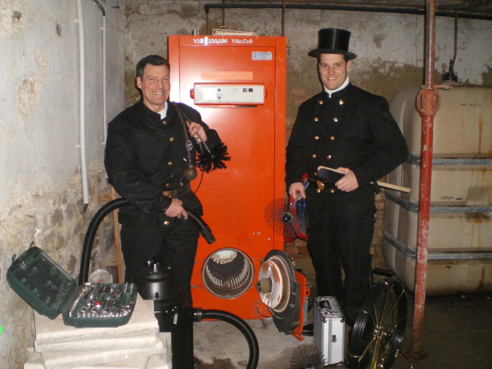 Deux ramoneurs alsaciens en tenue traditionnelle avec hérissons et matériel de ramonage dans une chaufferie