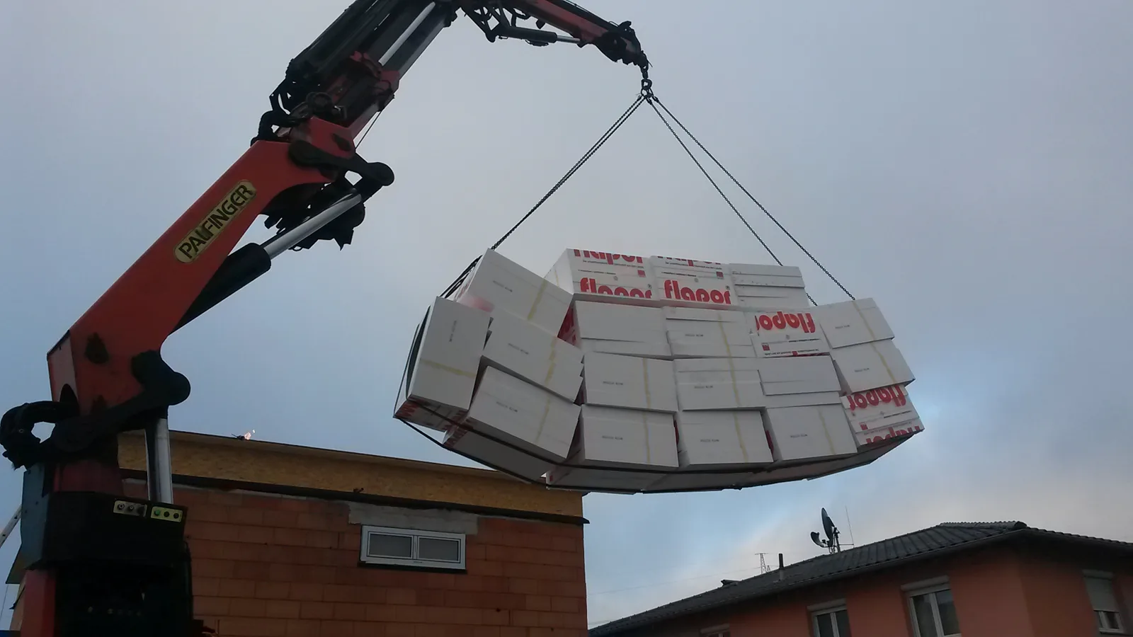 Grue de chantier levant une palette de panneaux isolants vers le toit plat d'une maison en rénovation