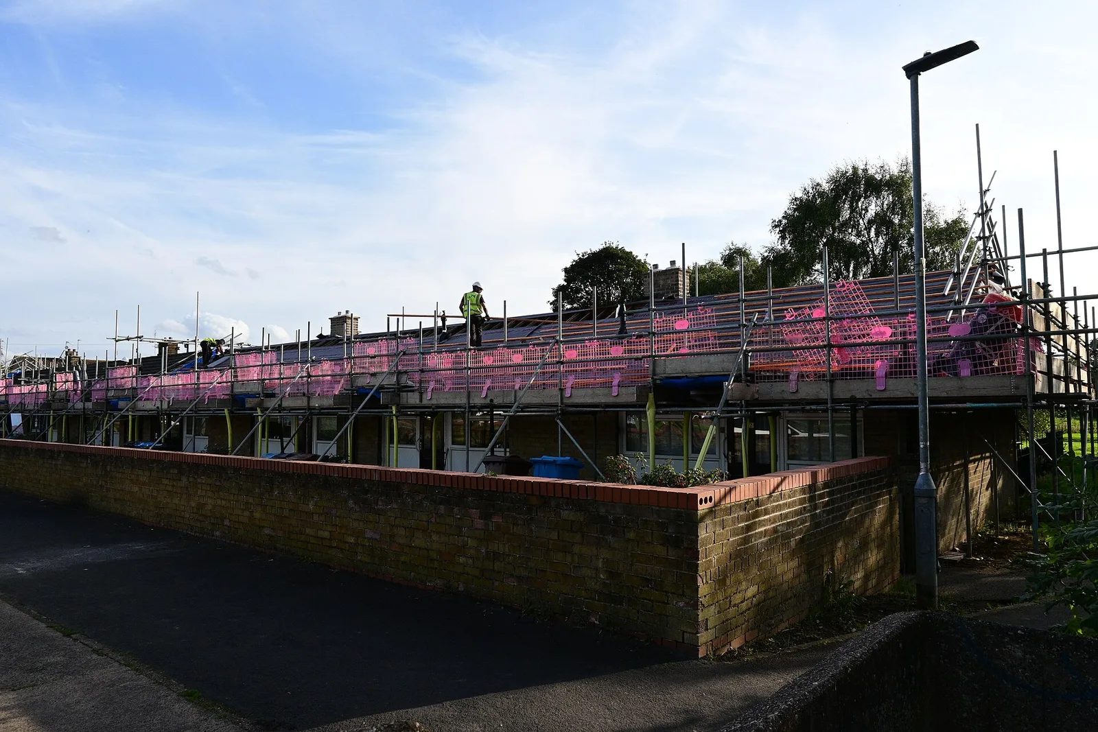 Rangée de maisons de plain-pied sous échafaudage pendant une rénovation de toiture, ouvrier en gilet jaune sur le toit et filets de protection roses