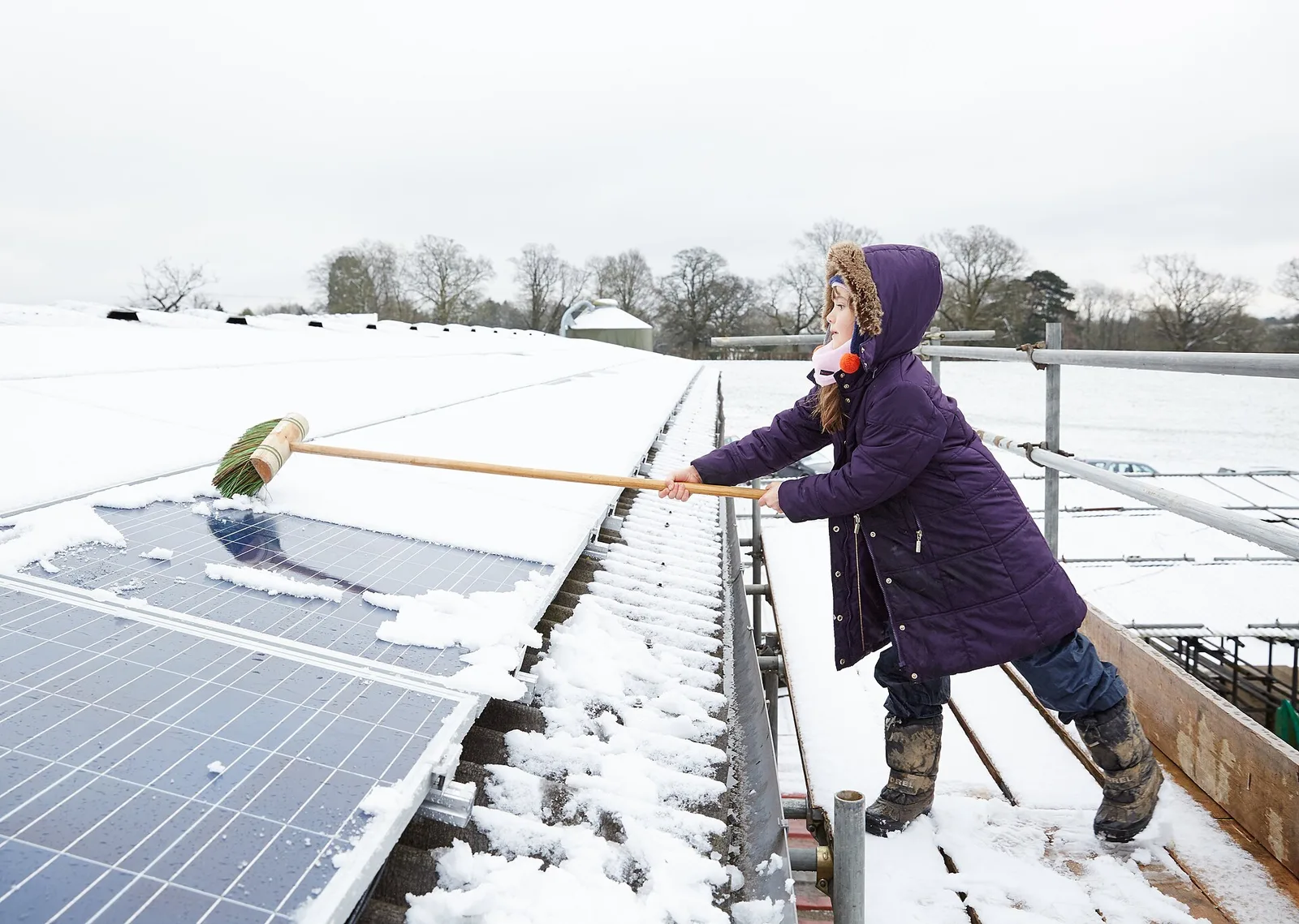 Enfant qui retire la neige de panneaux solaires avec un balai doux depuis un échafaudage muni d'un garde-corps, sans monter sur les modules