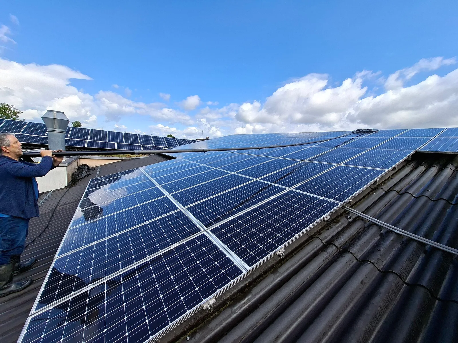 Homme debout sur un toit qui nettoie une rangée de panneaux solaires avec une longue perche télescopique munie d'une brosse alimentée en eau