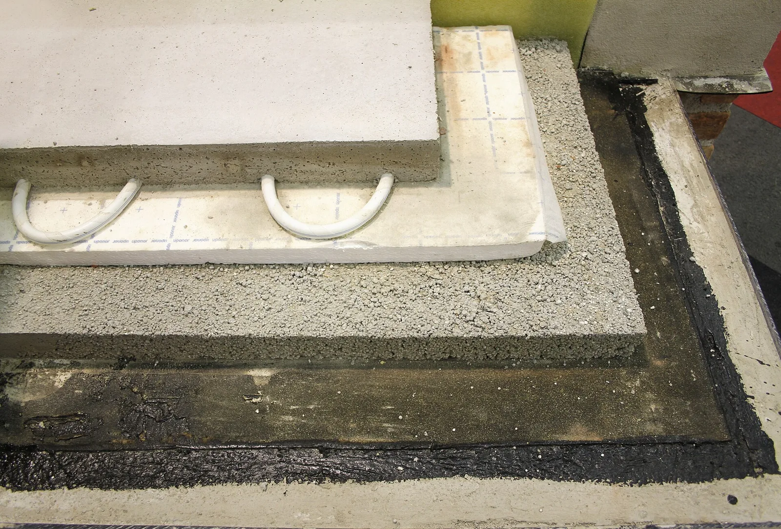Coupe d'un plancher chauffant : bitume d'étanchéité, couche de granulats, panneau isolant avec tubes blancs en U, chape béton posée dessus