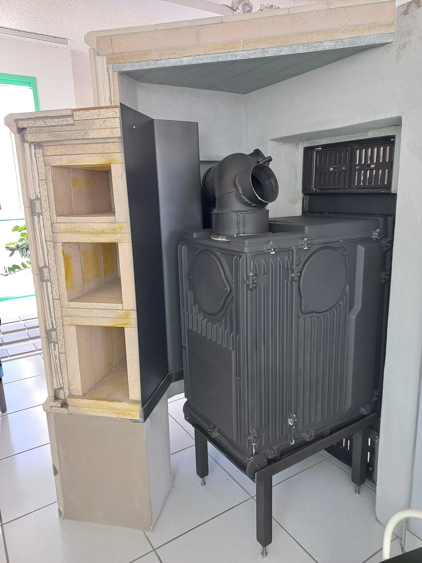 Poêle en faïence en cours de montage dans une maison autrichienne : foyer en fonte posé sur pieds, tuyau de raccordement, et à gauche une colonne de modules d'accumulation en chamotte empilés