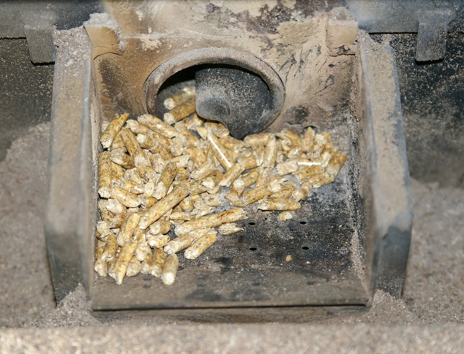 Creuset d'un poêle à granulés rempli de pellets, avec la vis d'alimentation au fond et les trous d'arrivée d'air dans la paroi, entouré de cendres
