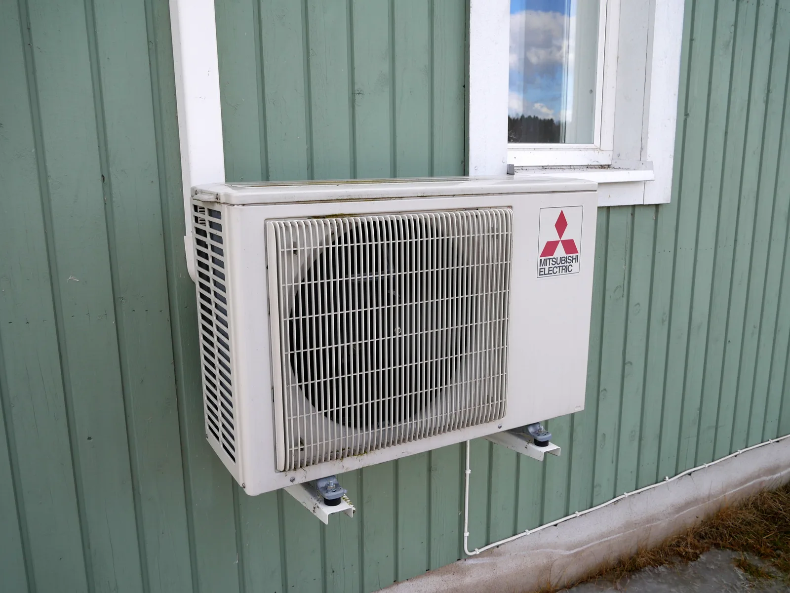 Unité extérieure de pompe à chaleur air-air mono-split fixée sur supports contre le bardage d'une maison, sous une fenêtre